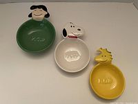 Three ceramic measuring cups featuring Peanuts characters Lucy, Snoopy, and Woodstock, with respective measurement markings 1/2 cup, 1 cup, and 1/4 cup visible.