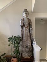 Front view of the full-height carved wooden Buddha statue on a wooden stand next to plants