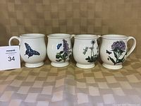 Photo of four Portmeirion ceramic mugs with various botanical and insect designs placed in a row on a patterned surface.