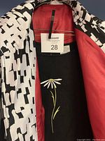 Open coat showing coral red lining with black and white geometric pattern exterior, over black dress with white daisy embroidery on front.