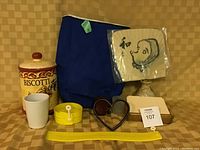 Photo showing biscotti jar, Japanese fan, yellow flower pot, two tea lights holders, index card file box, yellow saki jar, heart-shaped metal holder, insulated backpack all arranged on a surface with a tan backdrop.