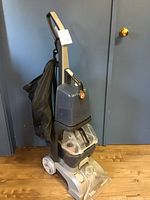 Hoover Turbo Scrub carpet shampooer standing upright showing front and side, with clear water tanks and black bag.
