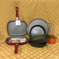 Flip Diamond red dual frying pan open showing non-stick interior surface with two compartments; two round 8 inch cake pans, rectangular loaf pan, cooling rack and small ceramic honey pot with orange and brown glaze.