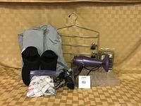 Photo showing all lot items arranged on beige surface against patterned beige background, including hair dryer, curling iron, slippers, hanger, bed sheet garters, make-up tray, shower hooks, and shoe holder.