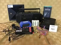 Photo of assorted electronic devices on a table showing radio cassette player, alarm clock, cassette player, TV antenna, USB car charger adaptors, tempered glass screen protector, cables, and power adapter.