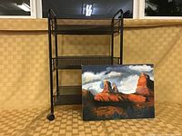 Black metal office trolley with three mesh shelves alongside a landscape painting showing red rock formations under a cloudy blue sky.