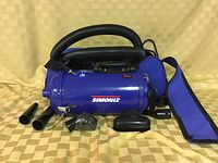 Blue Simoniz portable vacuum with black flexible hose, a blue shoulder strap, and multiple black attachments for different purposes arranged in front.