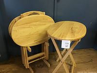 Multiple light wooden oval TV tables with foldable legs resting on the floor next to a wooden stand under dark wall background.