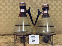 Two vintage clear glass coffee carafes with black handles and silver trim on black metal tea light warmers.