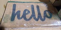 Rectangular natural coir doormat with black 'hello' script, packaged in clear plastic.