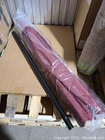 Photo of the wrapped red umbrella canopy with black pole laid on a wooden pallet and cardboard surface.