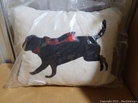 Photo of the front of the decorative pillow showing the black Labrador dog with a red scarf on a white background inside plastic packaging.