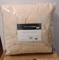 Front view of the cream-colored comforter inside clear plastic packaging, showing the Madison Park label with product name and size indication.