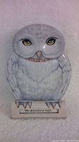 Close-up image of the Harry Potter Hedwig Owl tin bank, shows white and gray owl figure with realistic yellow eyes and base plaque saying 'The Adventures begin'