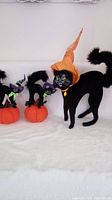 Lot of three Annalee black cat figurines on white background. One large cat with orange witch hat and necklace, two smaller cats on pumpkins with green hats.