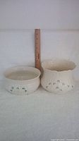 Two white porcelain Belleek pieces placed side by side with a wooden ruler behind them showing scale. One is a round shallow fruit bowl, the other a more textured planter with scalloped rim and shamrocks decoration.