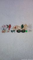 Photo showing a selection of Christmas-themed pins and earrings including sparkly cardinal pin, holly pin, silver bells earrings, snowflake pin, and two bulb pins.