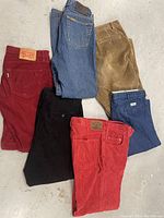 Six pairs of pants in various colors and brands on a floor showing back pocket and partial labels.