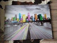 Large framed canvas print of a city skyline with buildings in multiple bright colors and highway light trails in foreground.