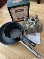 Lot including Hansel and Gretel bundt cake pan, black saucepan, box and booklet arranged on hardwood floor.