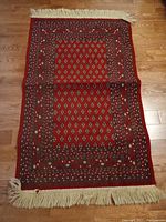 Full view of the small Persian style rug laid out on a wooden floor showing the red base with patterned design and cream fringes on short edges.