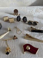 Photo showing a collection of vintage cufflinks, pins, tie clips, and beaver coin holder arranged on a white textured cloth. Items include various shapes and materials with a vintage look.