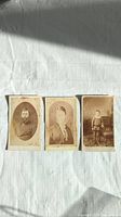 Three antique photographs laid out side by side on white fabric with sunlight casting shadows. Photos show portraits of a man, woman, and child. Moderate foxing and stains visible.
