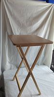 Photo showing a side view of one wooden TV table with X-shaped folding legs and a light wood rectangular top.