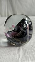 Rounded glass paperweight with black, pink, and gold abstract pattern inside, photographed on white surface showing the front side.