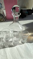 Full set of the clear glass decanter with stopper and three matching shot glasses displayed on a white surface with natural light highlighting etched designs.