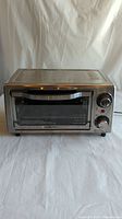 Front view of the Hamilton Beach toaster oven showing stainless steel body, glass door, and two control knobs with indicator light above.
