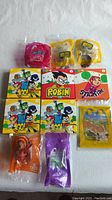 Eleven McDonald's Happy Meal toys displayed, five are Teen Titans Go! figures with colorful cardboard inserts, others include Trolls and Sing 2 figures, all in original packaging.
