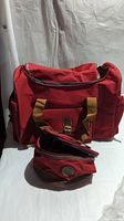 Red carry all duffel bag with brown handles and a matching toiletries bag in red with brown accents shown side by side.
