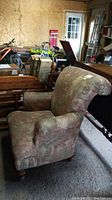 Front angled view of fabric upholstered armchair showing overall shape, wood legs, and wear on fabric with dusty environment background.