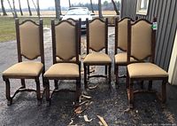 Front view of 6 carved wood dining chairs with upholstered tan seat and back, showing nailhead detail and carved feet, placed outside.