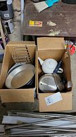 Two open cardboard boxes with assorted kitchenware on a workbench. Includes metal baking pans, white teapot, pot with handle, and metal baking tray holders.