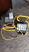 Two yellow treble flood work lights with black protective cages and attached power cords, shown on concrete floor.