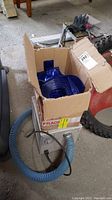 Box of pool parts with blue hoses coiled inside and a black pool pump placed next to the box.
