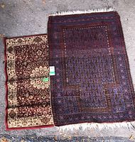 Two overlapping rugs outdoors on pavement, showing a dark blue and red patterned rug with damage to fringe on the left, and a beige and burgundy floral smaller rug underneath on the right.