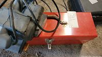 Red metal toolbox with latch on the floor with the belt sander's power cord draped over it, showing size and condition.