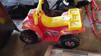 Front-left angled view of red and yellow ride-on toy ATV showing body, seat, steering handle, and large black wheels.