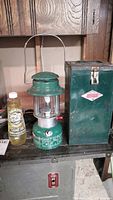 Full view of green Coleman Easi-Lite lantern next to metal case and bottle of lamp oil on shelf.