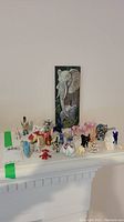 Photo showing entire collection of multiple elephant figurines arranged on mantle, featuring various materials including ceramic, glass, enameled brass, and a Dumbo figure. Ruanne Manning elephant art is propped upright behind them.