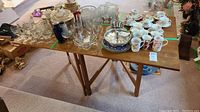 Top view of the solid mahogany folding table showing the surface with scratches, scuffs, and drop leaves extended. A variety of glassware and china are placed on the table for scale, not included in the lot.