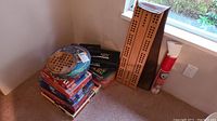 Stack of assorted board games and puzzles on the floor including a circular tin of Bongo game, several colorful boxed games stacked vertically, two cribbage boards, a rolled map or poster, and a large book.