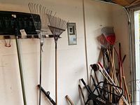 Wall mounted rack holding several garden tools including rakes and shovel, seen from a side angle showing handles and heads.