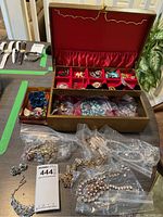 Photo showing gold-tone jewelry box with red velvet interior, containing various pins and jewelry pieces in compartments and a lower drawer with more jewelry in plastic bags lying in front.