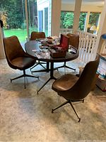 Four brown vinyl padded MCM style swivel dining chairs surrounding a woodgrain laminate dining table with various small items on the table.