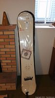 Full view of the snowboard leaning against a wall, wrapped in clear plastic. Shows graphics and length of the board.