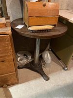 Round wood top table with a metal pedestal base, shown with a wooden box and brush on top. Located in a basement surroundings.
